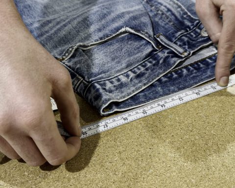 A close-up of hands measuring the waistband of a pair of blue jeans laid flat on a table using a tape measure, demonstrating how to measure the waist of jeans accurately.
