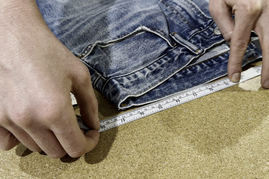 A close-up of hands measuring the waistband of a pair of blue jeans laid flat on a table using a tape measure, demonstrating how to measure the waist of jeans accurately.