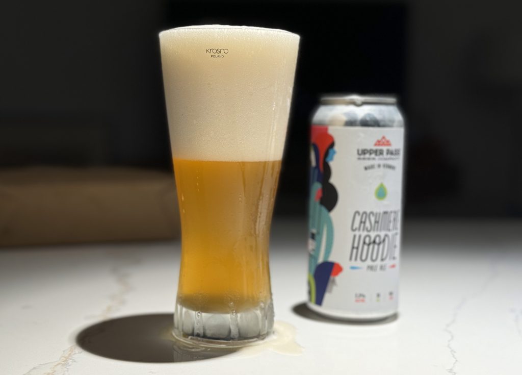 An ice cold, tall Krosno beer glass filled with Cashmere Hoodie Pale Ale by Upper Pass Beer Company with a thick, foamy head next to the can captures the hazy, golden-yellow appearance, topped with a thick, foamy head.