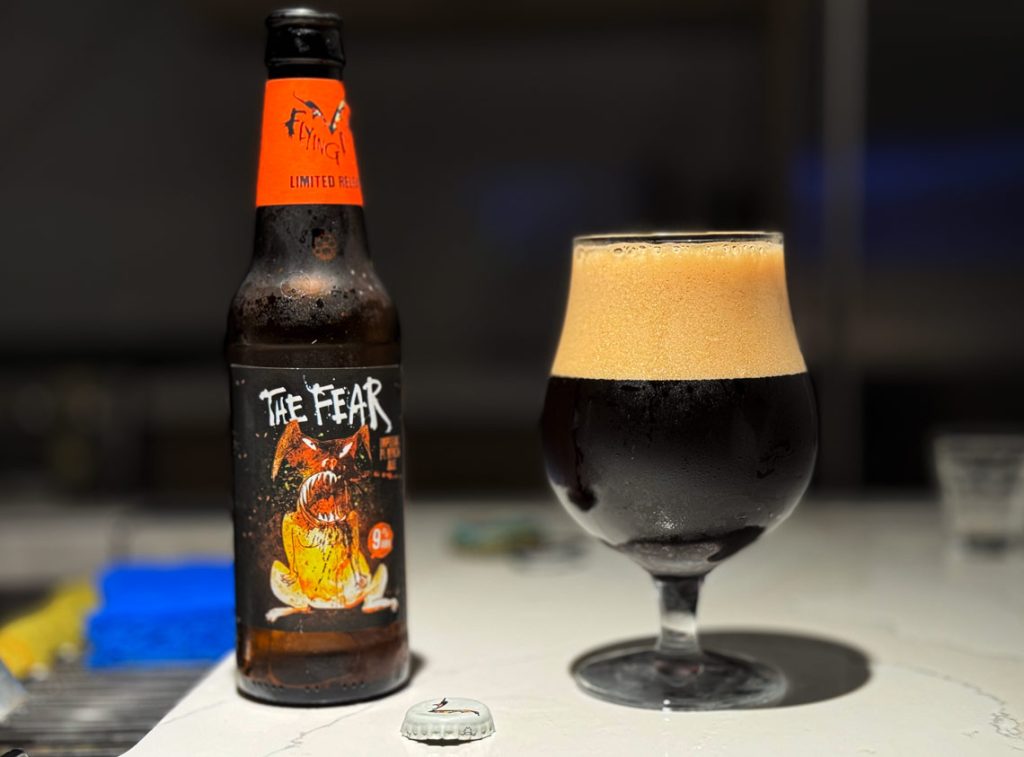 12-ounce bottle of Flying Dog Brewery's The Fear Imperial Pumpkin Ale next to a glass filled with the dark beer, showcased on a countertop with a blurry background emphasizing the beer's creamy head and rich color.