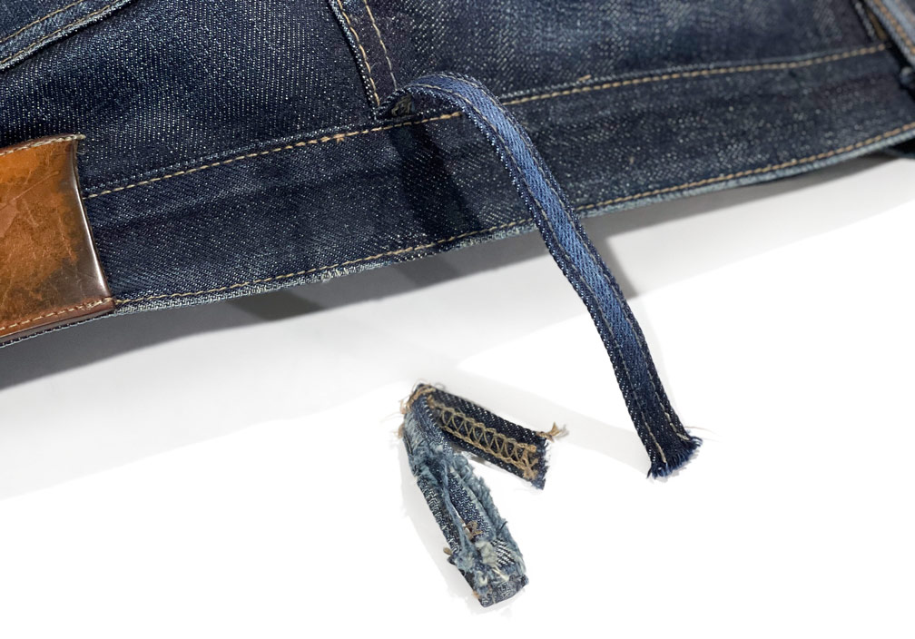 A pair of jeans showing a newly attached replacement belt loop beside the original worn, detached loop, highlighting professional repair work by the Williamsburg Garment Company's belt loop repair service.