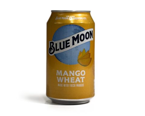 A can of Blue Moon Mango Wheat beer, photographed on a white background, for a Denimbmc.com blog review. The can features bright yellow-orange packaging with a mango illustration.