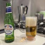 A bottle of Peroni Nastro Azzurro next to a glass mug of poured beer with a foamy head on a kitchen counter, with a stove and espresso maker in the background.