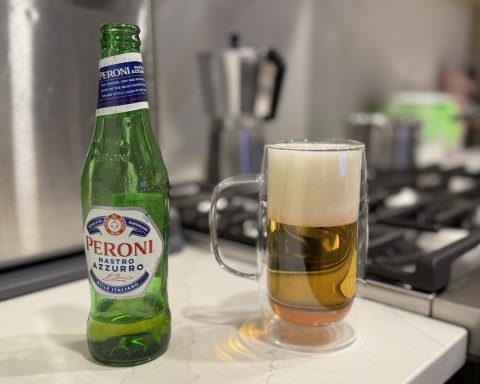 A bottle of Peroni Nastro Azzurro next to a glass mug of poured beer with a foamy head on a kitchen counter, with a stove and espresso maker in the background.