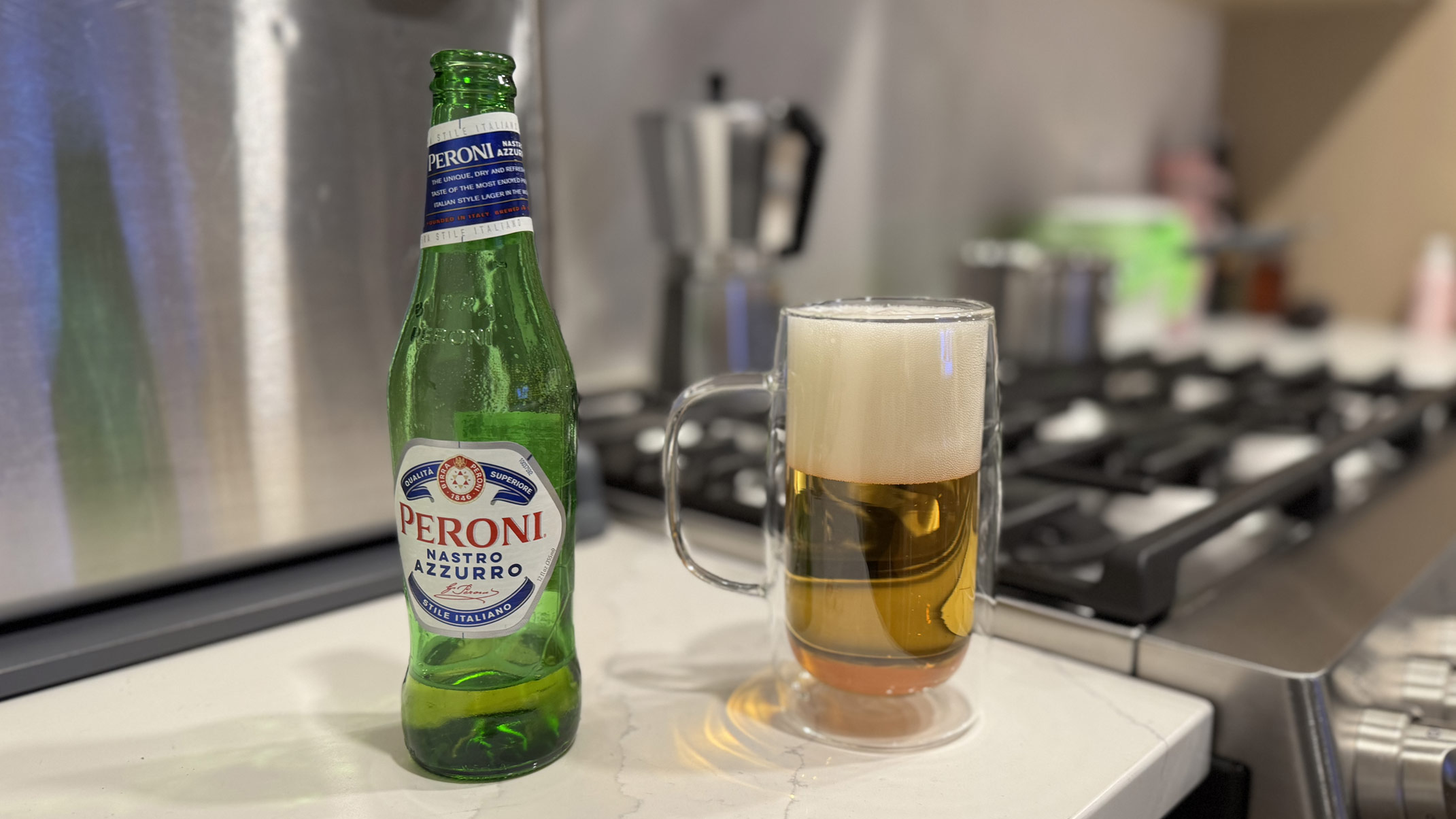A bottle of Peroni Nastro Azzurro next to a glass mug of poured beer with a foamy head on a kitchen counter, with a stove and espresso maker in the background.